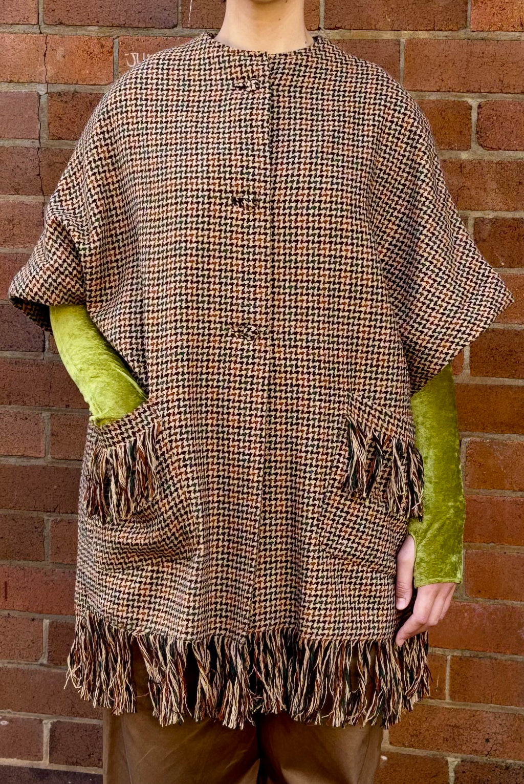 Person wearing a brown plaid poncho with green sleeves against a brick wall.