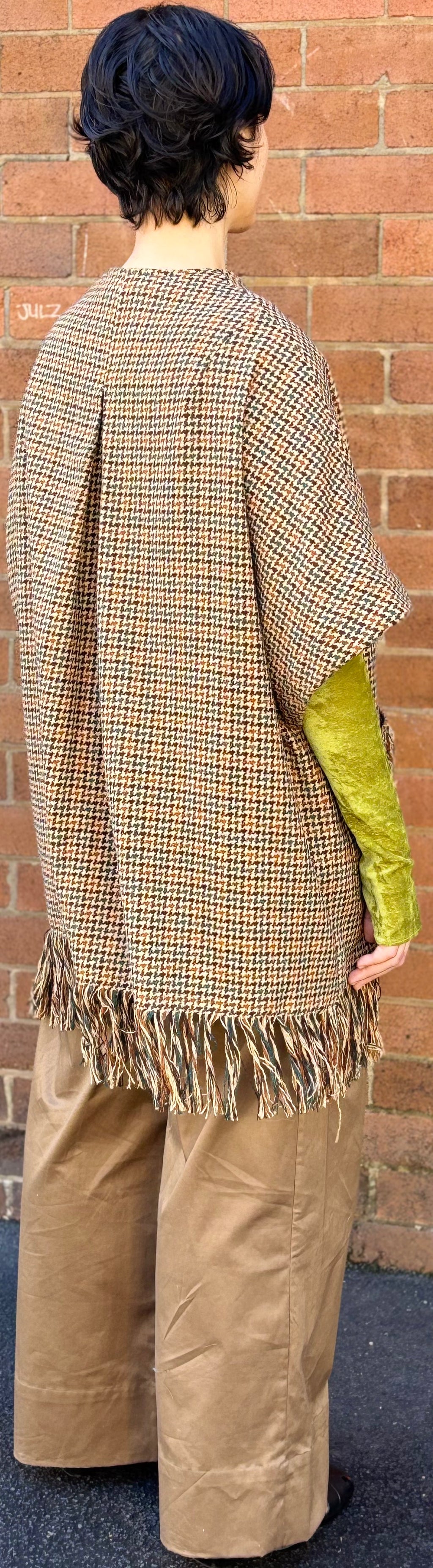 Person wearing a plaid shawl and beige pants against a brick wall.