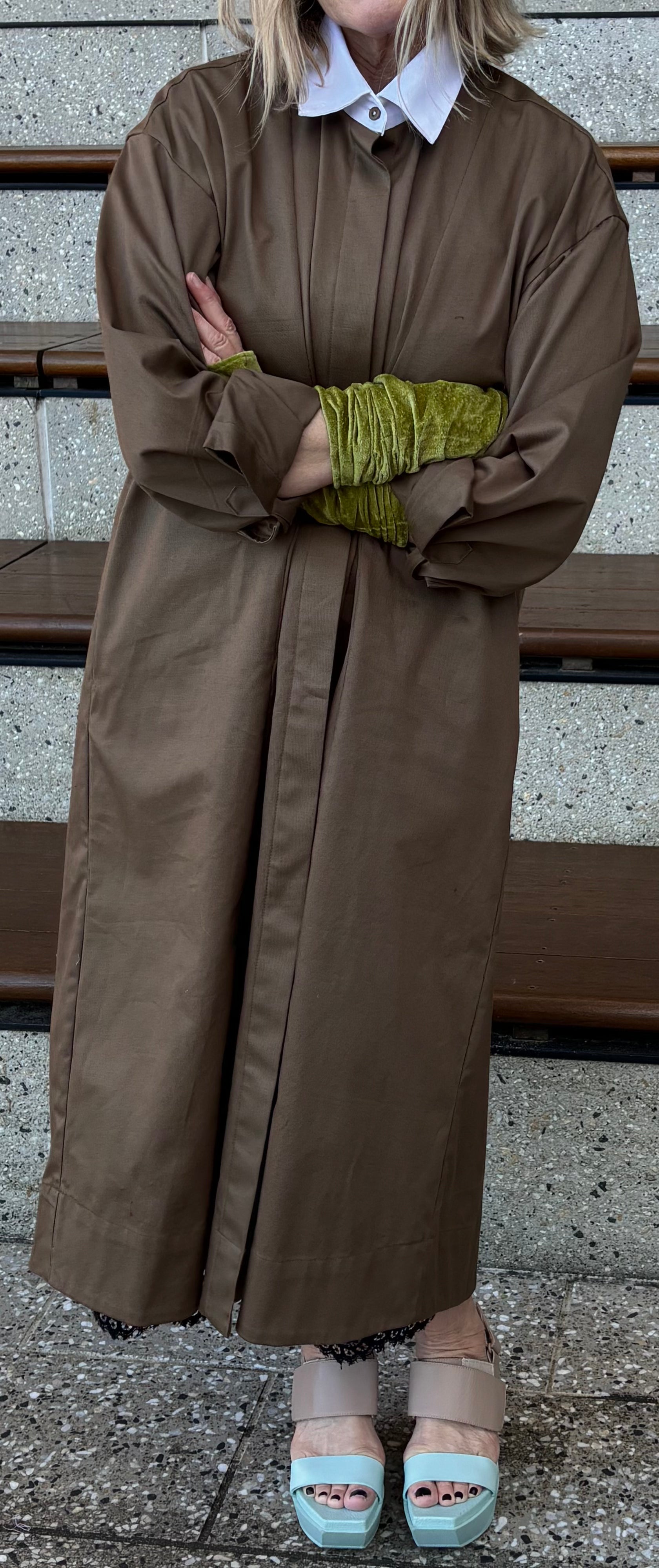Person wearing a long brown coat with green gloves, standing on a stone surface.